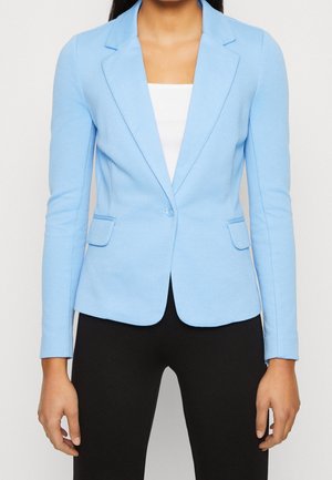 Frau trägt einen taillierten hellblauen Blazer über einem weißen Top und schwarzen Hosen, die Hände entspannt an den Seiten.