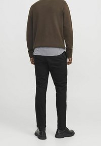 Brązowy sweter o fakturze, noszony na czarnej koszuli w białe paski. Czarne spodnie i czarne buty, widziane od tyłu.