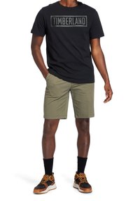 Camiseta negra de manga corta con un logo gris de "Timberland", combinada con pantalones cortos verdes olivo y zapatillas deportivas negras y naranjas.