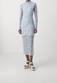 Abito azzurro chiaro a maniche lunghe con silhouette aderente, caratterizzato da un motivo a catena giallo e grigio a spirale. Indossato con tacchi alti bianchi.