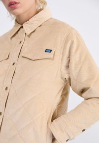 Beige kordfløyels skjortejakke med diamant quilt mønster, knapp front, to brystlommer, og KAVU logo lapp på venstre lomme.