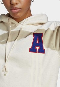 Kreativ huvtröja i krämfärgad med broderad "A"-logotyp i blått och rött, snörning i huven och vertikala ränder på tyget. Mjuk textur, avslappnad design.