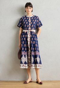 Mørkeblå midikjole med blomstermønster, korte ermer og et hvitt belte. Kjolen har trykt splitt og en kremfarget knapplukking.