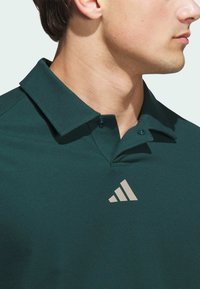 Dark green polo shirt with a textured weave, two buttons on the collar, and a light gray Adidas logo on the chest.