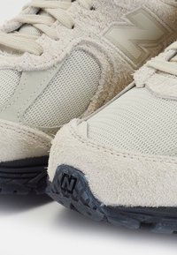 Gros plan de baskets beige en suède et mesh avec semelles noires, équipées de panneaux texturés et de lacets, mettant en avant le logo de la marque sur le bout et le côté.
