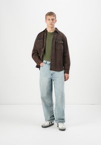 Brun denim button-up skjorte, grønn strikket t-skjorte under, lyseblå vide bukser og svart og hvit joggesko. Enkle, uformelle klær.