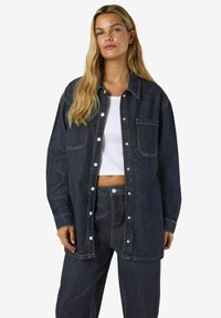 Jeans srajca temno modre barve, z udobnim krojem, sprednjim zapiranjem na gumbe in žepom na prsih. Gladek tekstura z vidnimi detajli šivov.