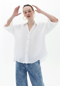 Camicia bianca in tessuto con colletto, parzialmente aperta e con spacchi laterali, abbinata a jeans in denim azzurri a vita alta.