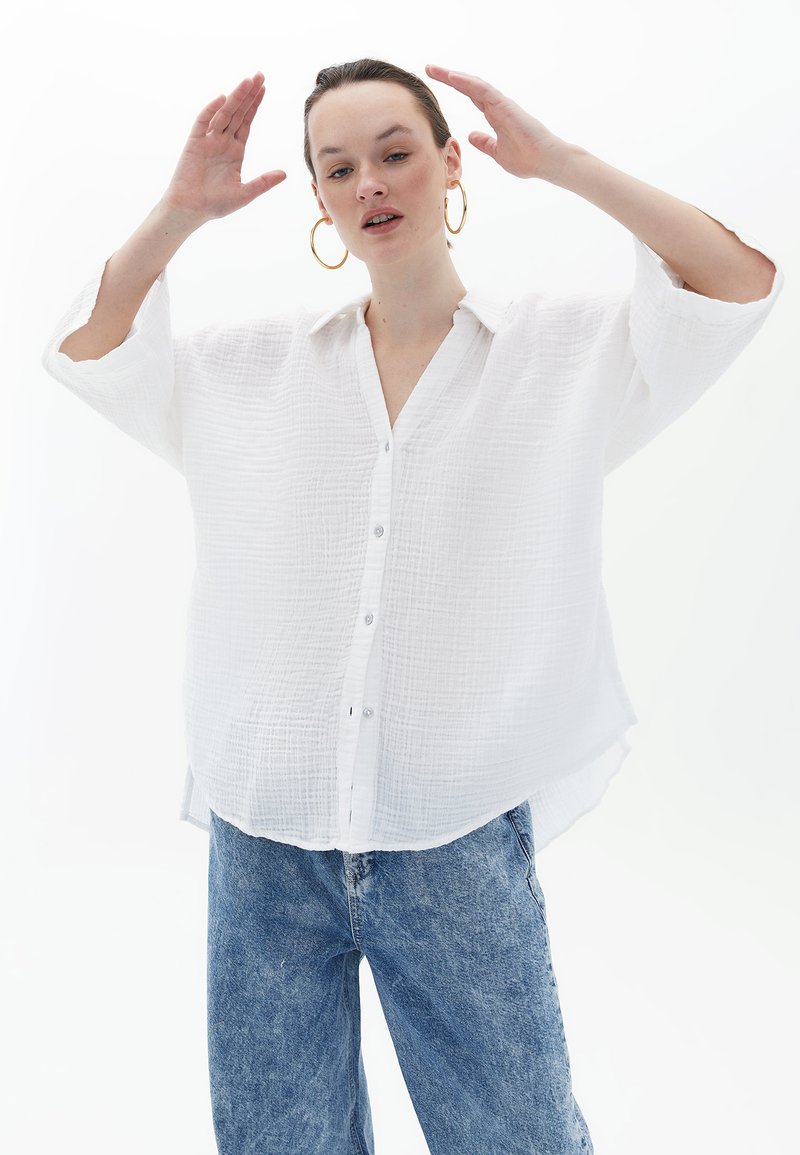 Camicia bianca in tessuto con colletto, parzialmente aperta e con spacchi laterali, abbinata a jeans in denim azzurri a vita alta.