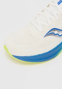 Weiße Sportschuh mit Mesh-Obermaterial, blauen und grünen Akzenten, ausgeprägter texturierter Sohle und einem Logo mit einem Wellenmuster.