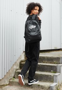 Zaino nero con logo Vans, realizzato in tessuto, forma rettangolare, spallacci regolabili e tasca frontale. Indossato da una persona mentre sale le scale.