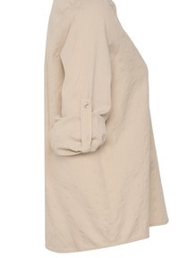 Chemise longue beige avec manches retroussées maintenues par une patte boutonnée, vue de profil sur un fond blanc uni.
