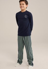 Mornarsko modra majica z dolgimi rokavi s grafiko slušalk, v kombinaciji z zelenimi in mornarsko modrimi prugastimi hlačami. Gladek material, sproščen kroj. Bele nogavice.