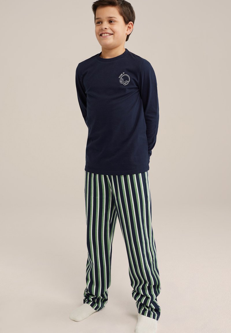 Mornarsko modra majica z dolgimi rokavi s grafiko slušalk, v kombinaciji z zelenimi in mornarsko modrimi prugastimi hlačami. Gladek material, sproščen kroj. Bele nogavice.