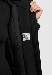 Zwarte pufferjack met een textuurstof, met een close-up van een label met tekst. Het interieur is glad en glanzend.