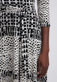 Robe à motif noir et blanc avec ceinture nouée à la taille, manches trois-quarts, portée par une personne à la peau foncée, main reposant sur le côté.