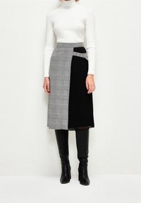 Woman wearing a black and gray plaid midi skirt with an attached belt, white ribbed turtleneck, and black knee-high boots, standing against white wall.
