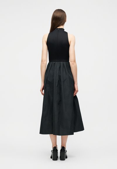 Femme debout de dos portant une robe midi noire sans manches avec une jupe froncée et des chaussures à talons noires, sur un fond uni.