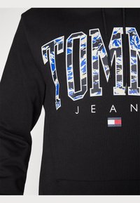 Sudadera negra con la palabra "TOMMY" en un estampado de camuflaje azul y blanco. Incluye "JEANS" en texto blanco y un pequeño logo de bandera. Material suave, mangas largas.