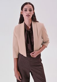 Beige cropped blazer met een gladde textuur, brede revers en driekwart mouwen, gecombineerd met een donkerbruine blouse met een strik.