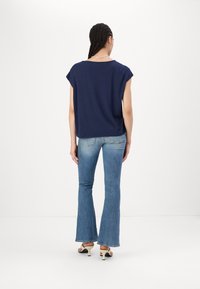 Marineblå kortermet topp med avslappet passform, sammen med flagrende lyseblå jeans og hæler, sett fra baksiden.