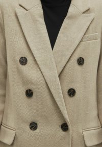 Beige fiskbensmönstrad blazer med dubbelknäppning, svarta knappar, slagkragar och en bröstficka. Tjock tygkänsla.