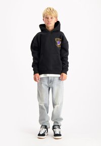 Zwarte hoodie met een voorzak en geborduurd ontwerp, gecombineerd met lichtblauwe versleten jeans. Gecombineerd met zwart-witte sneakers.