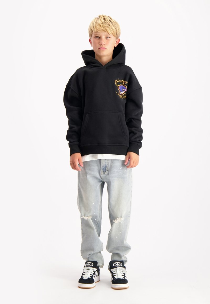 Zwarte hoodie met een voorzak en geborduurd ontwerp, gecombineerd met lichtblauwe versleten jeans. Gecombineerd met zwart-witte sneakers.