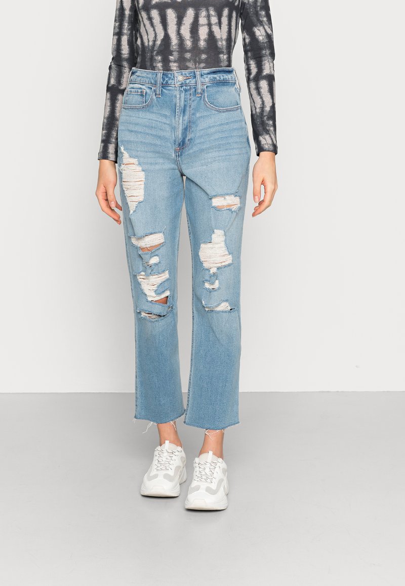 Jeans de cintura alta, azul claro, com rasgões desgastados e bainhas desfiadas, combinados com ténis brancos. Feitos de denim com uma textura suave.