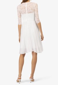 Robe blanche avec un corsage en dentelle ajusté, des manches trois-quarts en dentelle et une jupe plissée. Fermeture éclair au dos, texture douce.