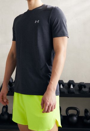 Mężczyzna w czarnej koszulce Under Armour z wzorem w kratkę i jasnych żółtych sportowych szortach na siłowni, w tle kettlebells i hantle.