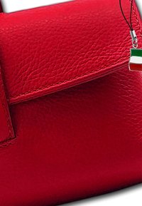 Rode leren handtas met een gestructureerde afwerking, voorzien van een klep aan de voorkant en een bedel van de Italiaanse vlag die aan een zwarte draad hangt.