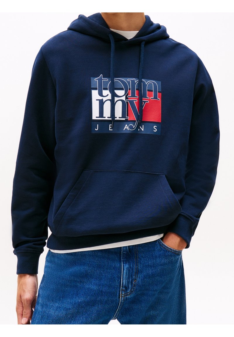 Homme portant un sweat à capuche bleu marine avec le logo "Tommy Jeans" en blanc, rouge et bleu, associé à un jean en denim bleu, main dans la poche.