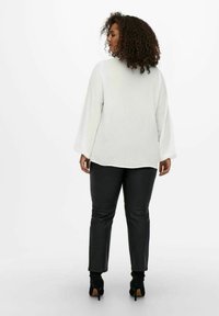 Blusa blanca de manga larga con un corte relajado, combinada con pantalones de imitación piel negros. El conjunto presenta mangas fluidas y un escote redondo.