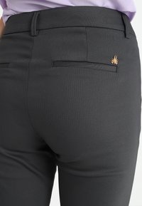 Zwarte op maat gemaakte broek met een gladde textuur, voorzien van achterzakken en een klein gouden logo-accent aan de rechterzijde.