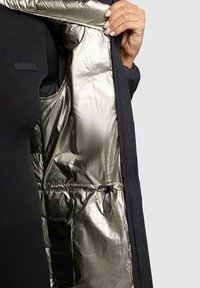 Veste en duvet noire non zippée, révélant une doublure métallique argentée brillante. Présente une texture extérieure matelassée et un détail de cordon de serrage ajustable.