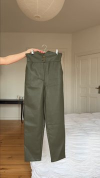 Pantalon en cuir vert olive taille haute avec double boutons dorés, suspendu à un cintre tenu par une personne dans une chambre minimaliste.