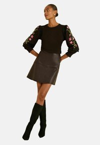 Fekete, hosszú ujjú felső virágmintás hímzéssel és fodros részletekkel, egy testhez simuló fekete bőr miniszoknyával és térd fölé érő fekete velúr csizmával párosítva.