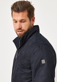 Marineblauer gesteppter Mantel mit hohem Kragen und Reißverschluss. Verfügt über einen strukturierten Stoff und ein Logo-Tag auf der linken Schulter.