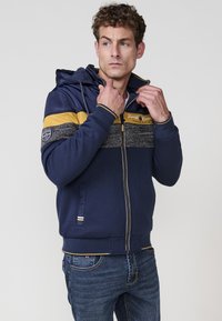 Sudadera con cremallera de color azul marino con acentos a rayas amarillas y grises. Presenta una capucha forrada de felpa y bolsillos laterales, confeccionada con un tejido suave y texturizado.