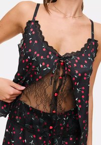 Top en satin noir avec imprimé cerises rouges, accents en dentelle, bretelles spaghetti réglables et une superposition en dentelle avec un détail à nouer.