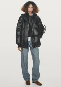 Osoba s kudrnatými vlasy, oblečená v černé péřové bundě, volných modrých džínách, hnědých botách, držící černou tašku na jednobarevném pozadí.