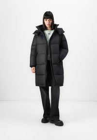 Woman wearing a long black puffer coat, light green sweatshirt, black pants, and black chunky shoes, standing against a white background.