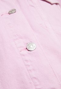 Camisa rosa con una tela texturizada, que presenta un botón plateado redondo marcado con "TOMMY HILFIGER" y detalles de costura visibles.
