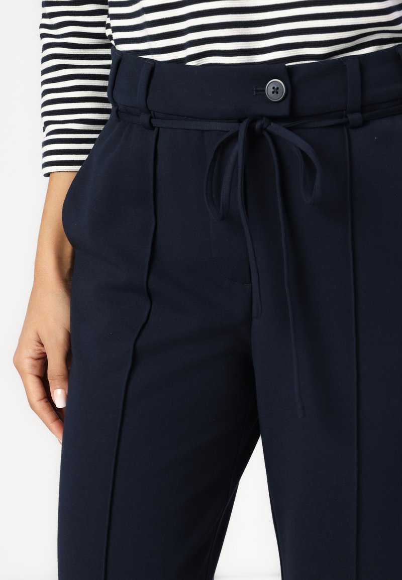 Granatowe spodnie z wysokim stanem, z paskiem do związania i fałdami z przodu. Materiał wydaje się gładki z lekkim streczem. Widoczny detal z zapięciem na guzik.