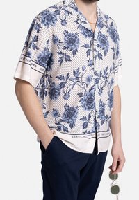 Man in een beige overhemd met korte mouwen met een blauw bloemmotief en zwarte stippen, navy broek, met heldere zonnebril in de rechterhand.