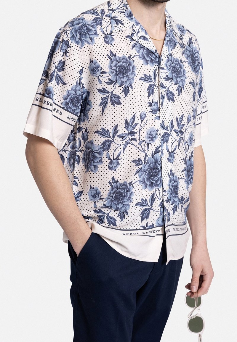 Man in een beige overhemd met korte mouwen met een blauw bloemmotief en zwarte stippen, navy broek, met heldere zonnebril in de rechterhand.