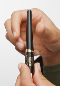 Eye-liner noir avec un design effilé, des accents dorés et un texte noir sur l'étiquette. Tenue entre des doigts contre un fond blanc.