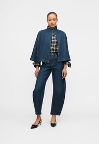 Γυναίκα στέκεται φορώντας κάπα από denim με κουμπιά τύπου snap, πουκάμισο καρό, τζιν ψηλόμεσο και μαύρες μυτερές γόβες kitten heels σε λευκό φόντο.