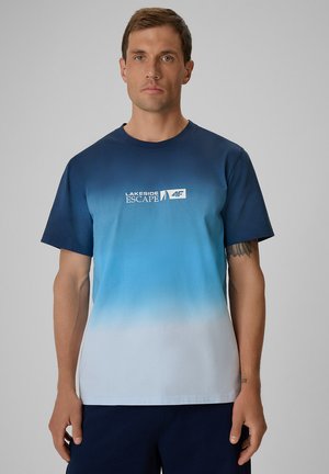 Mężczyzna z krótkimi brązowymi włosami w t-shircie o gradiencie z niebieskiego do białego z napisem "Lakeside Escape" i logo, stojący na jednolitym tle.